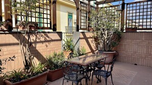 An outdoor patio with a glass table and metal chairs, surrounded by potted plants and a brick wall. - Hotel Suite Ares (Naples)