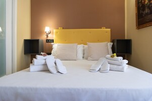 Habitación de hotel con cama, ropa de cama blanca, cabecera amarilla, dos altavoces fijados a la pared y un cuadro en la pared.
