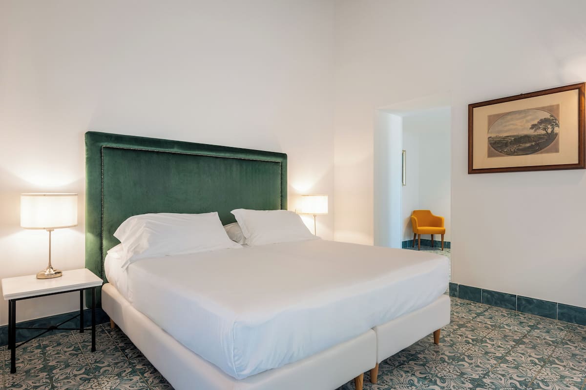 Una cama bien hecha con un cabecero tapizado en verde, dos mesitas de noche con lámparas, un cuadro enmarcado en la pared y un suelo con diseño.