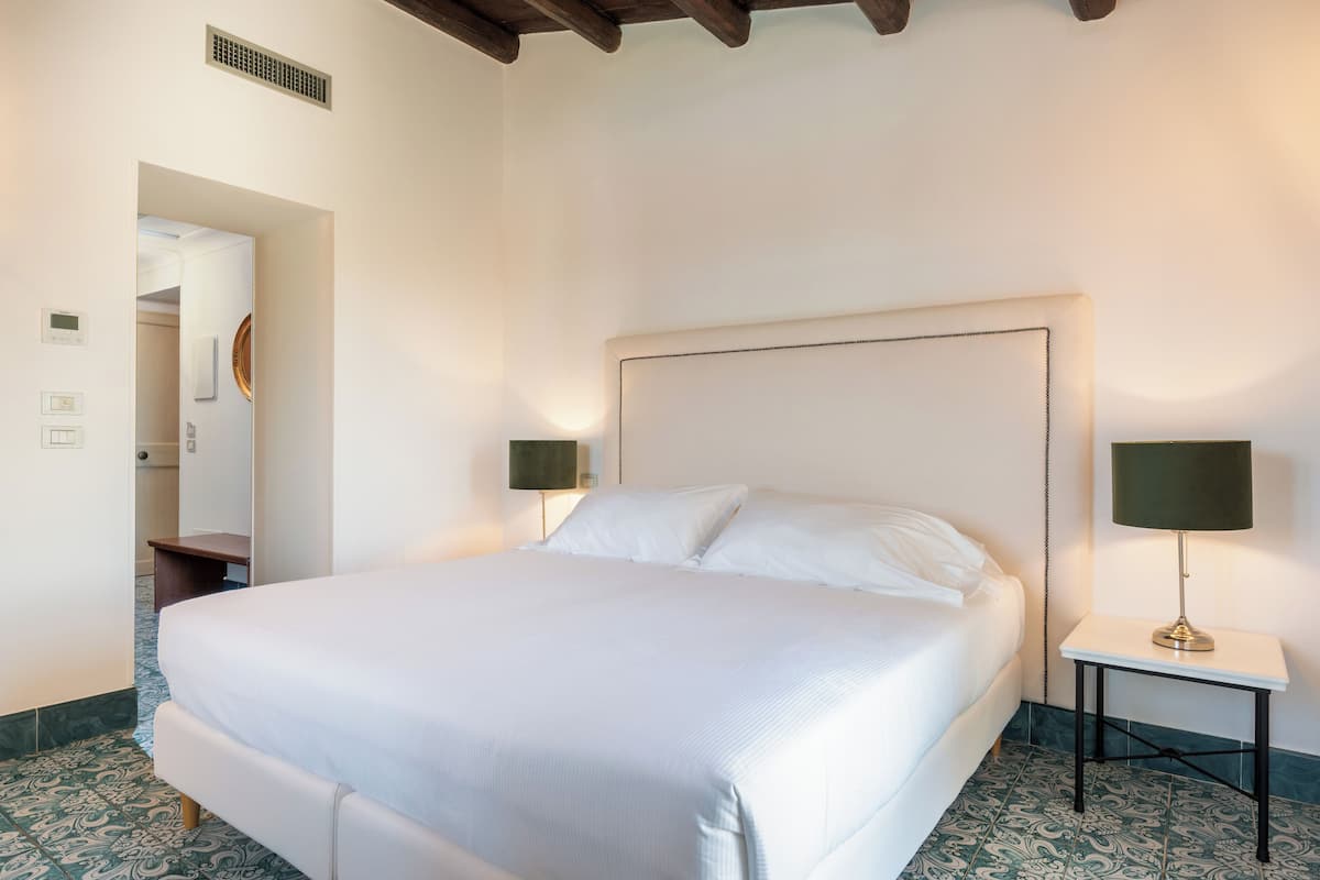 Una cama bien hecha en una habitación de hotel con un cabecero grande, dos mesitas de noche con lámparas y un suelo estampado.
