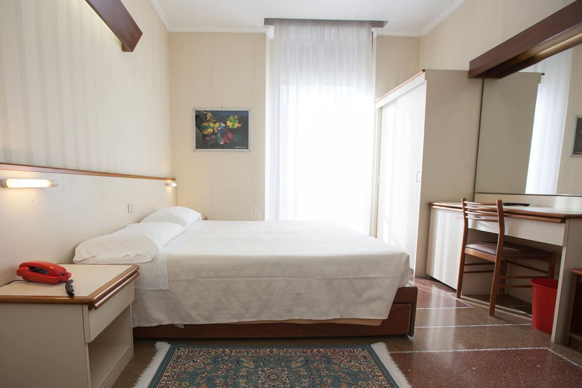 A hotel room with a bed, bedside table, desk, chair, and a large window with curtains.