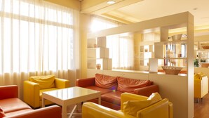 A modern living room with yellow and orange furniture, large windows with sheer curtains, and a kitchen area visible through a glass partition. - Hotel Regent (Pescara)