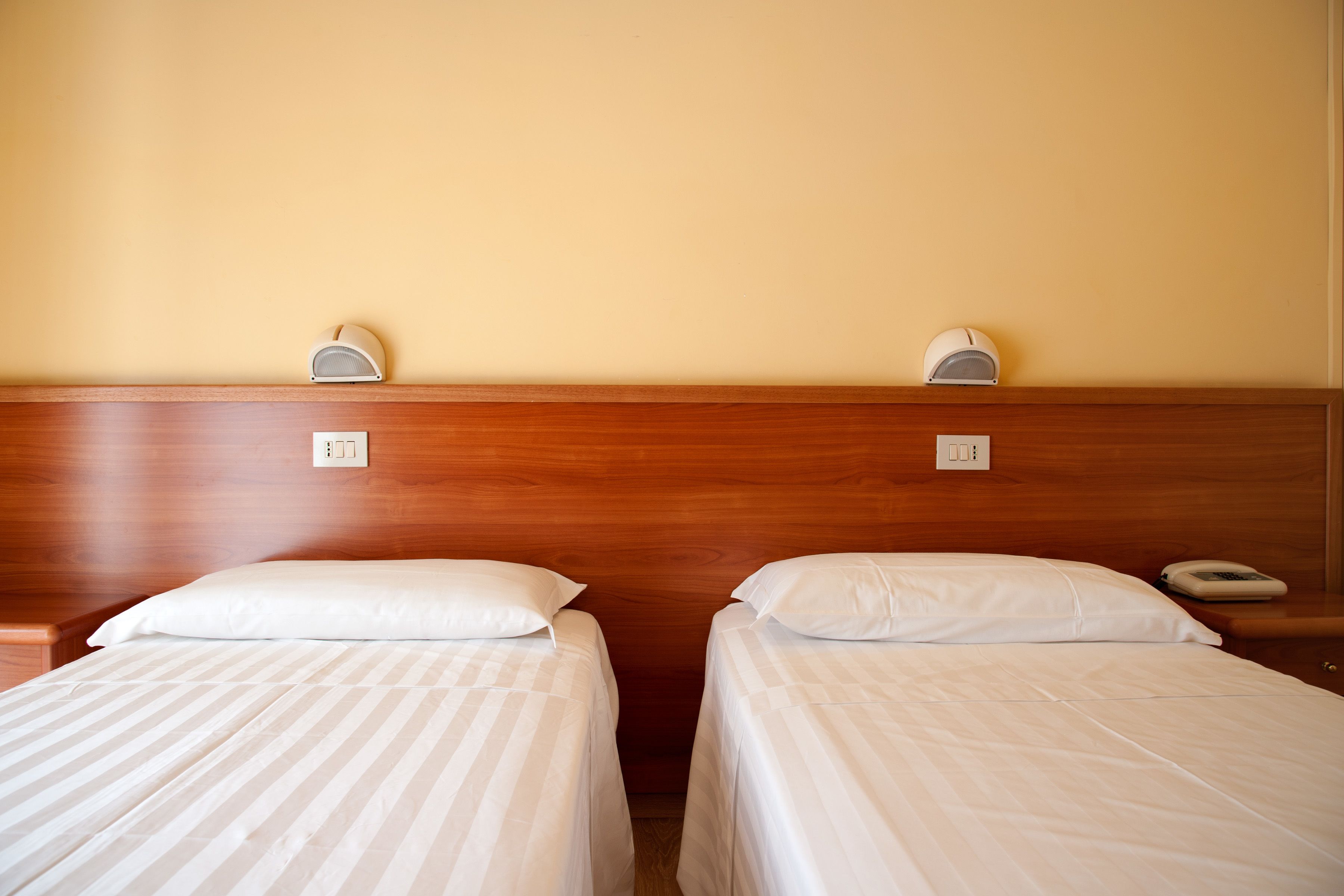 Camera d'albergo con due letti singoli, poggiatesta in legno, comodini e lampade a parete.