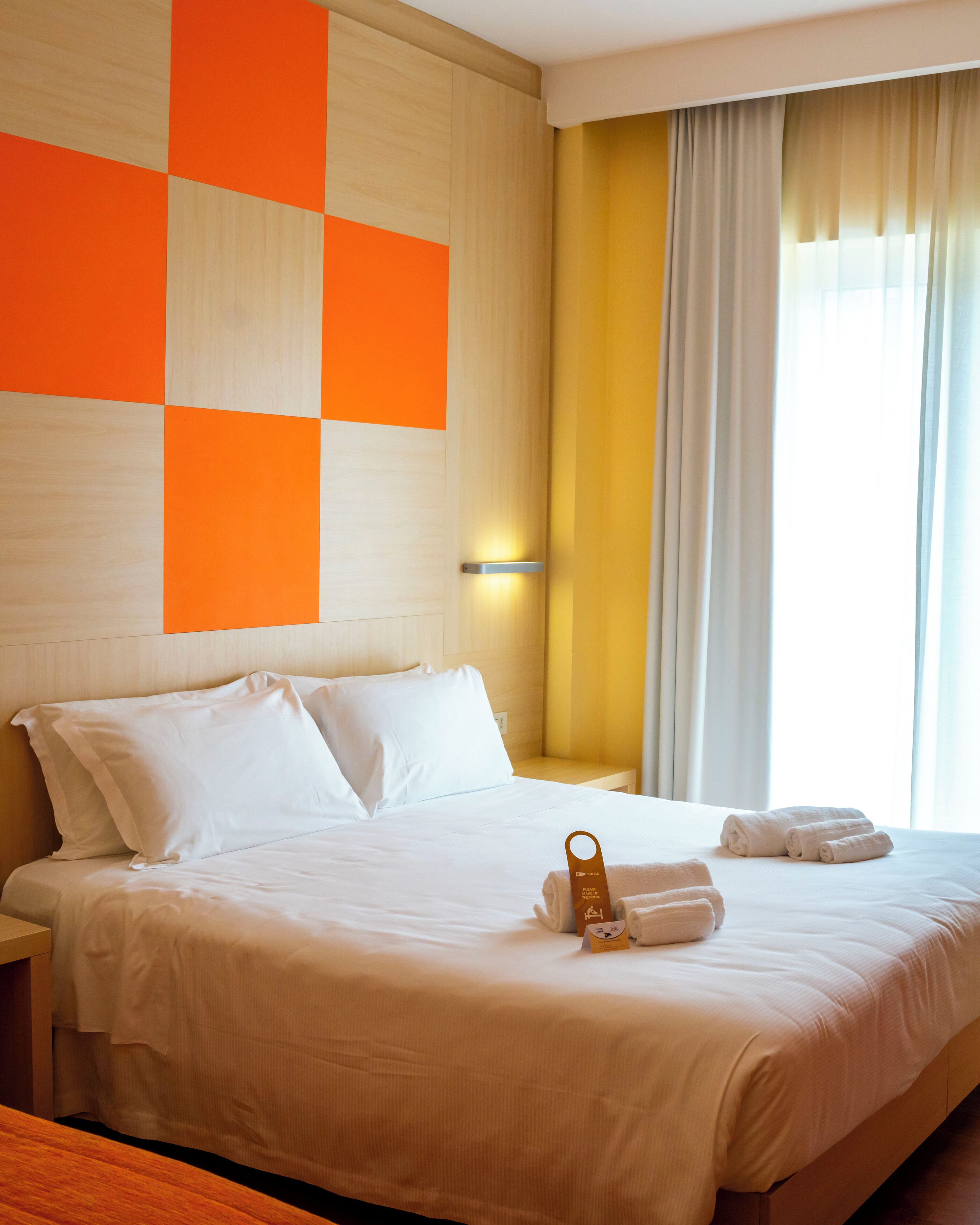 A hotel room with a bed, pillows, a bedside table, a wall-mounted light, and a window with curtains.