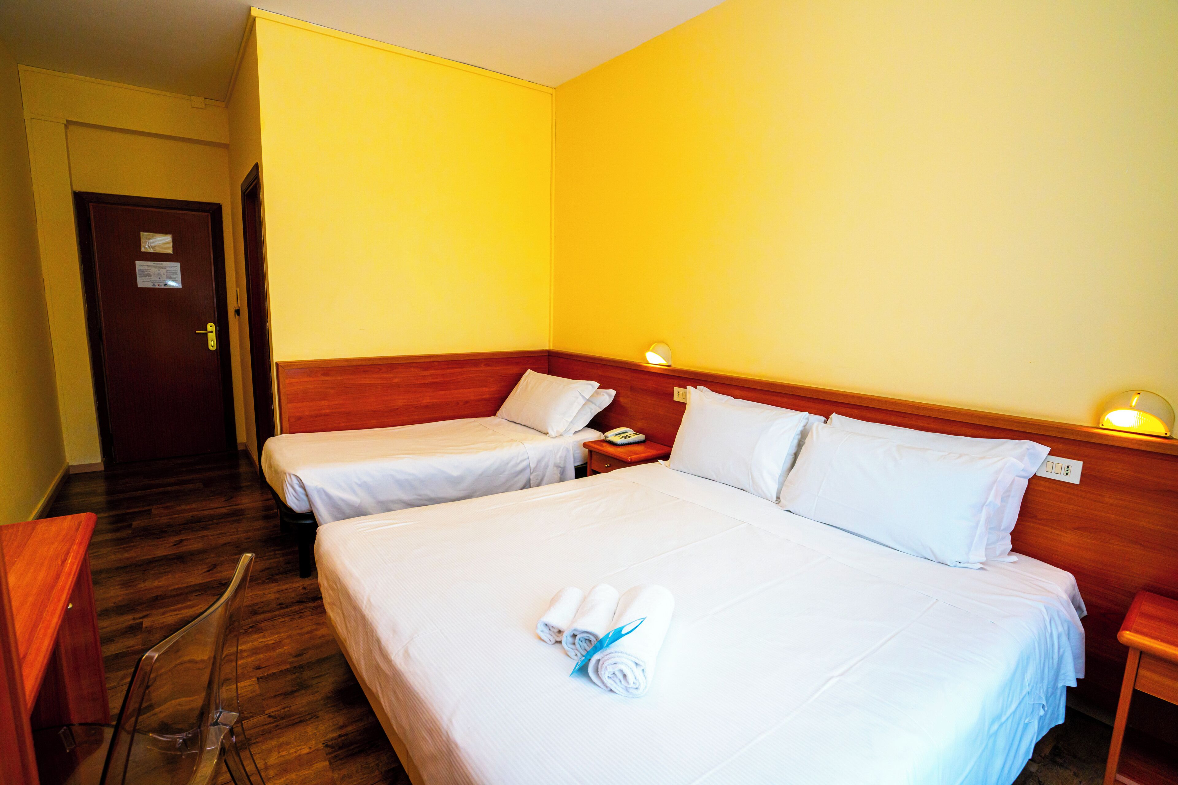 Habitación de hotel con dos camas individuales, cabecero de madera, mesita de noche con lámpara y puerta con un cartel.