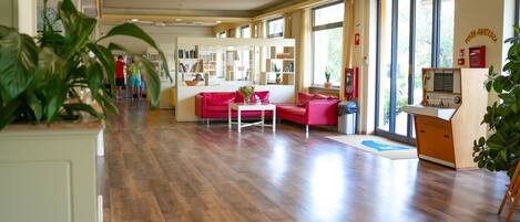 A spacious room with a wooden floor, a red sofa, a white table, and a large window with a view of greenery.