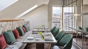 A dining area with a high table set for four, green chairs, a sofa with patterned cushions, and a view of a cityscape through large windows. - Vista Verona (Verona)