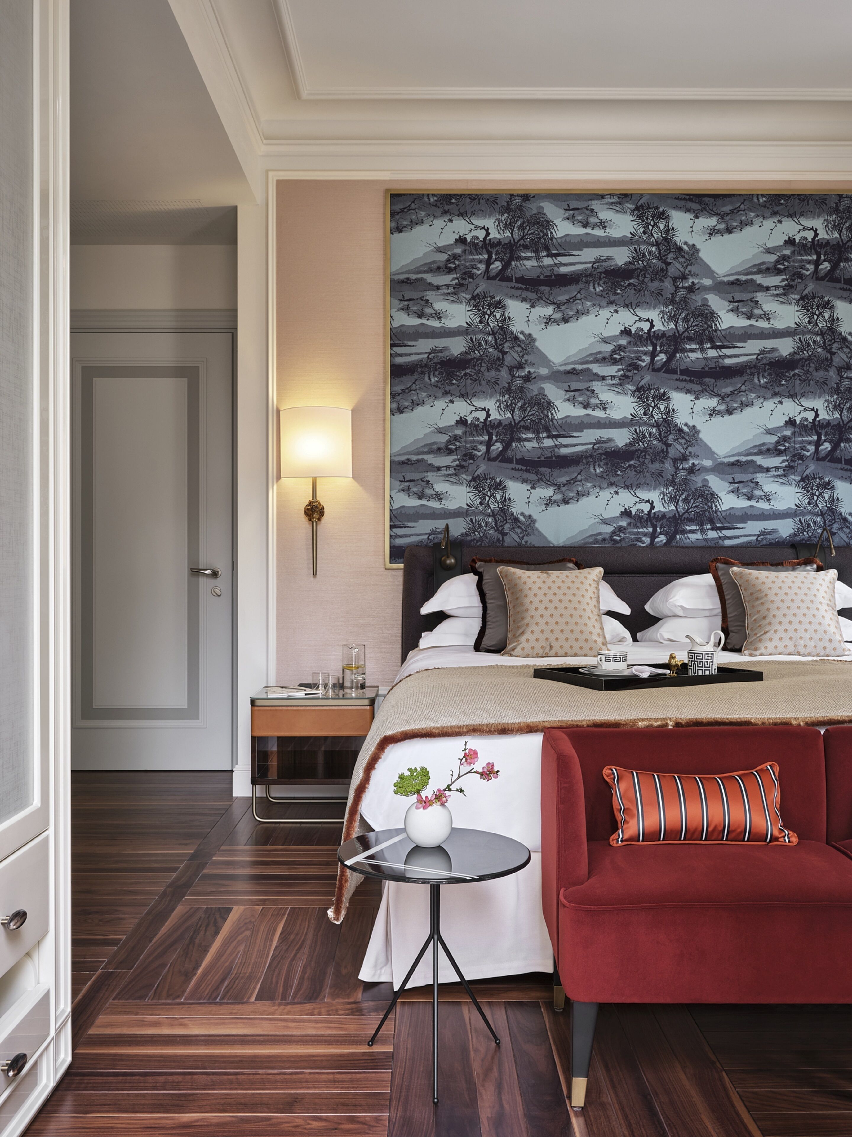 A hotel room with a bed, a nightstand, a table with a vase, and a red sofa.