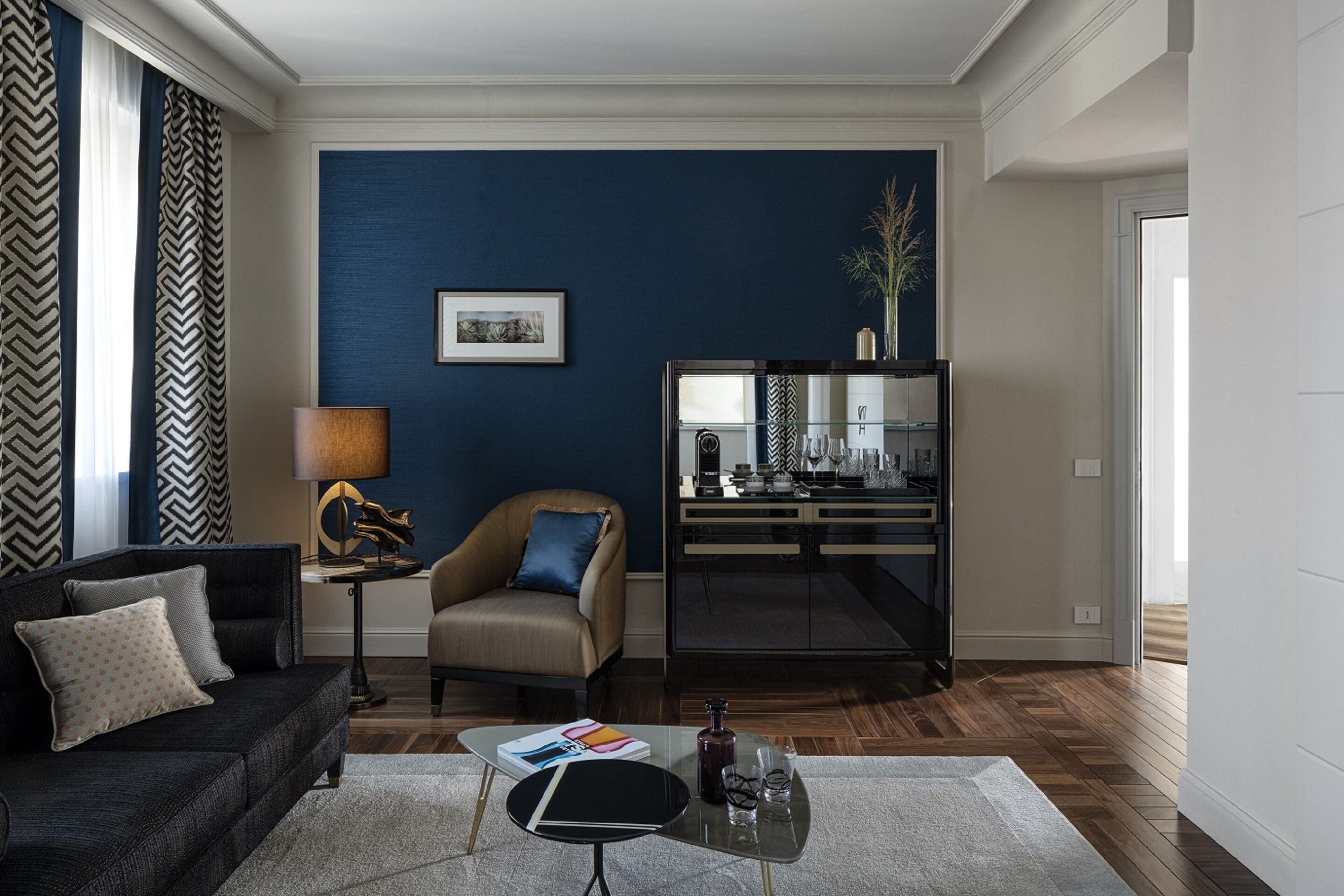 Una sala de estar moderna con una pared de acento azul oscuro, un sofá, una silla, una mesa de centro de vidrio y un aparador.