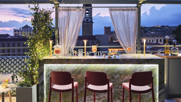 A rooftop bar with a view of a historic tower, two chairs, and a bar counter with drinks and a candle. - Vista Verona (Verona)
