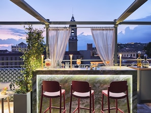 A rooftop bar with a view of a historic tower, two chairs, and a bar counter with drinks and a candle.
