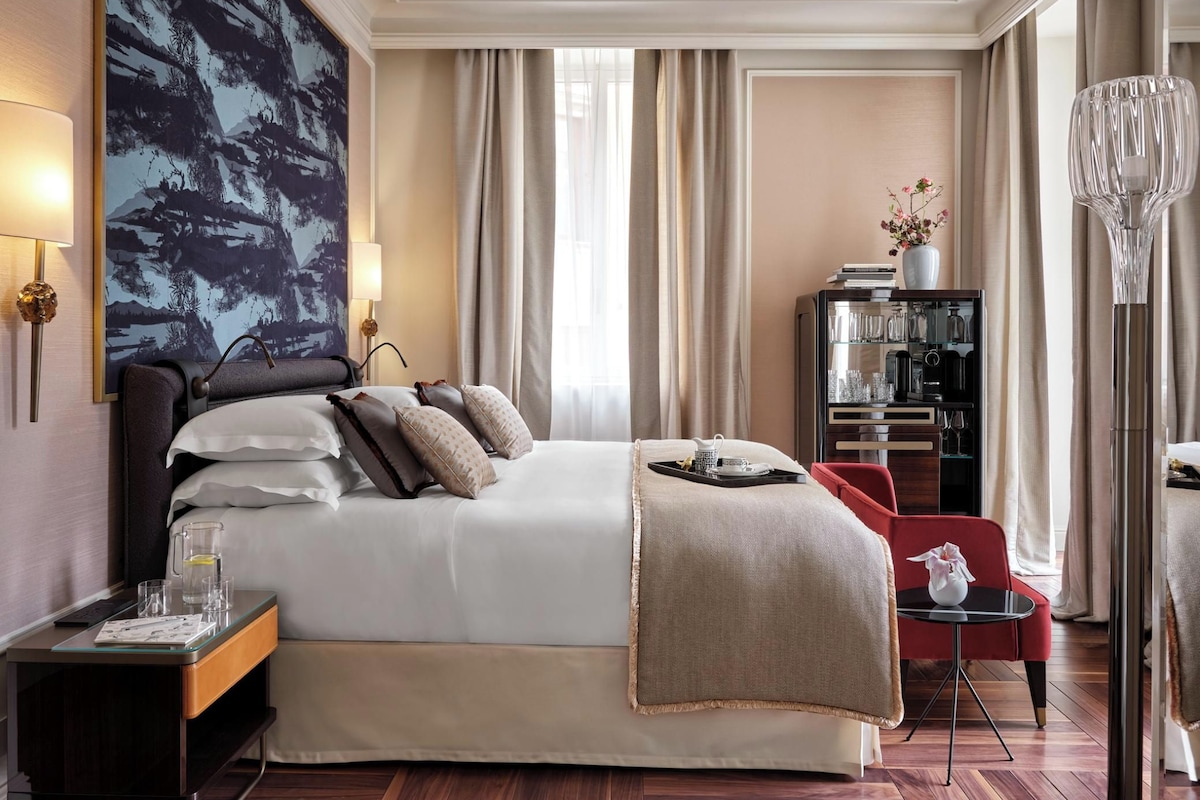 Una habitación de hotel con una cama grande, una mesita de noche con lámpara, una pequeña mesa con un florero y una estantería.