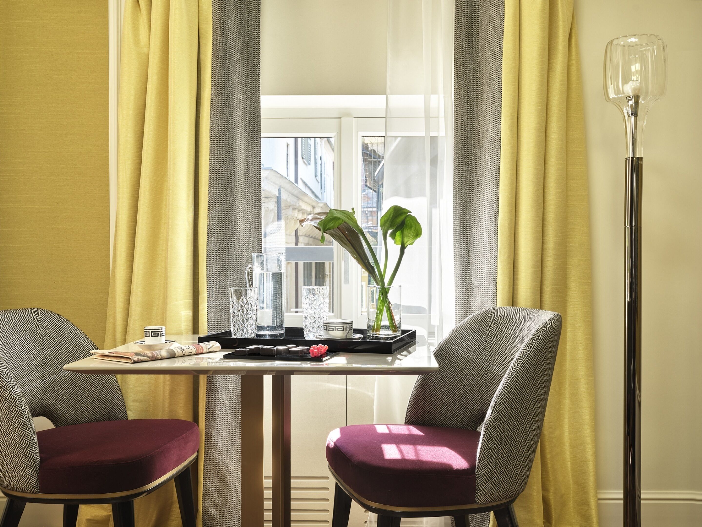 A hotel room with a dining table, two patterned chairs, a vase with a plant, and a floor lamp.