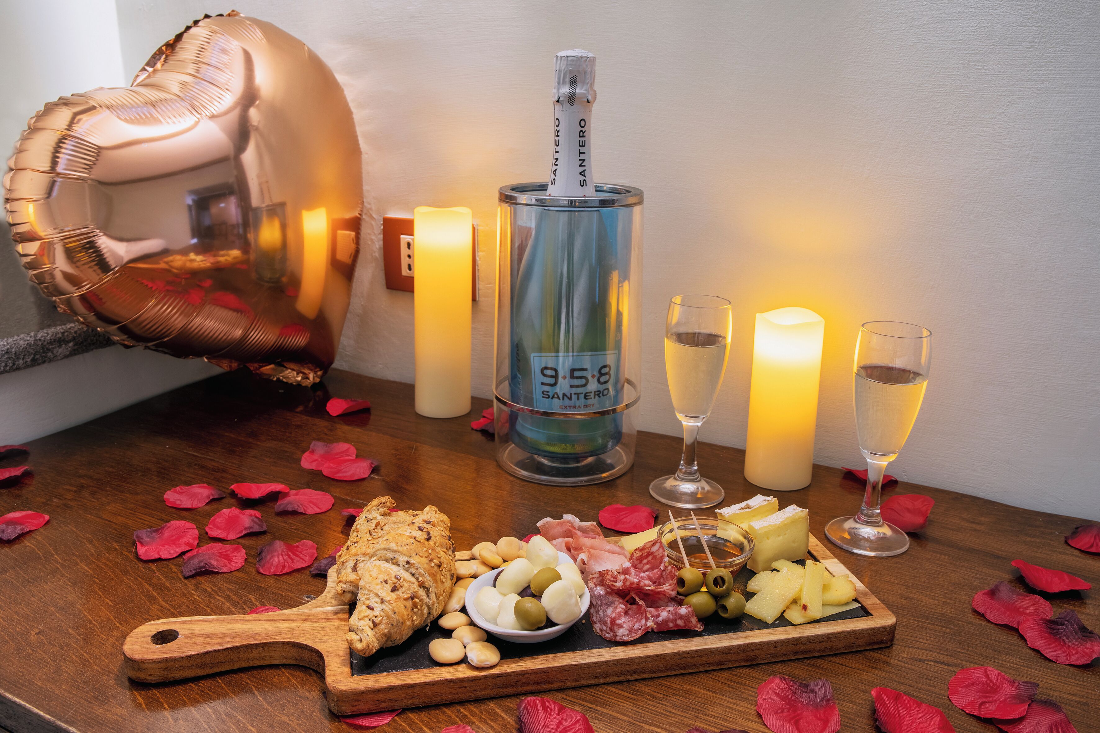 Un tavolo con una bottiglia di vino, bicchieri, un tagliere di formaggi, olive e salumi, circondato da candele e petali di rosa.