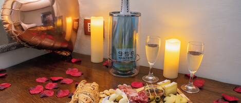 A table with a bottle of wine, glasses, a wooden board with cheese, olives, and meats, surrounded by candles and rose petals.