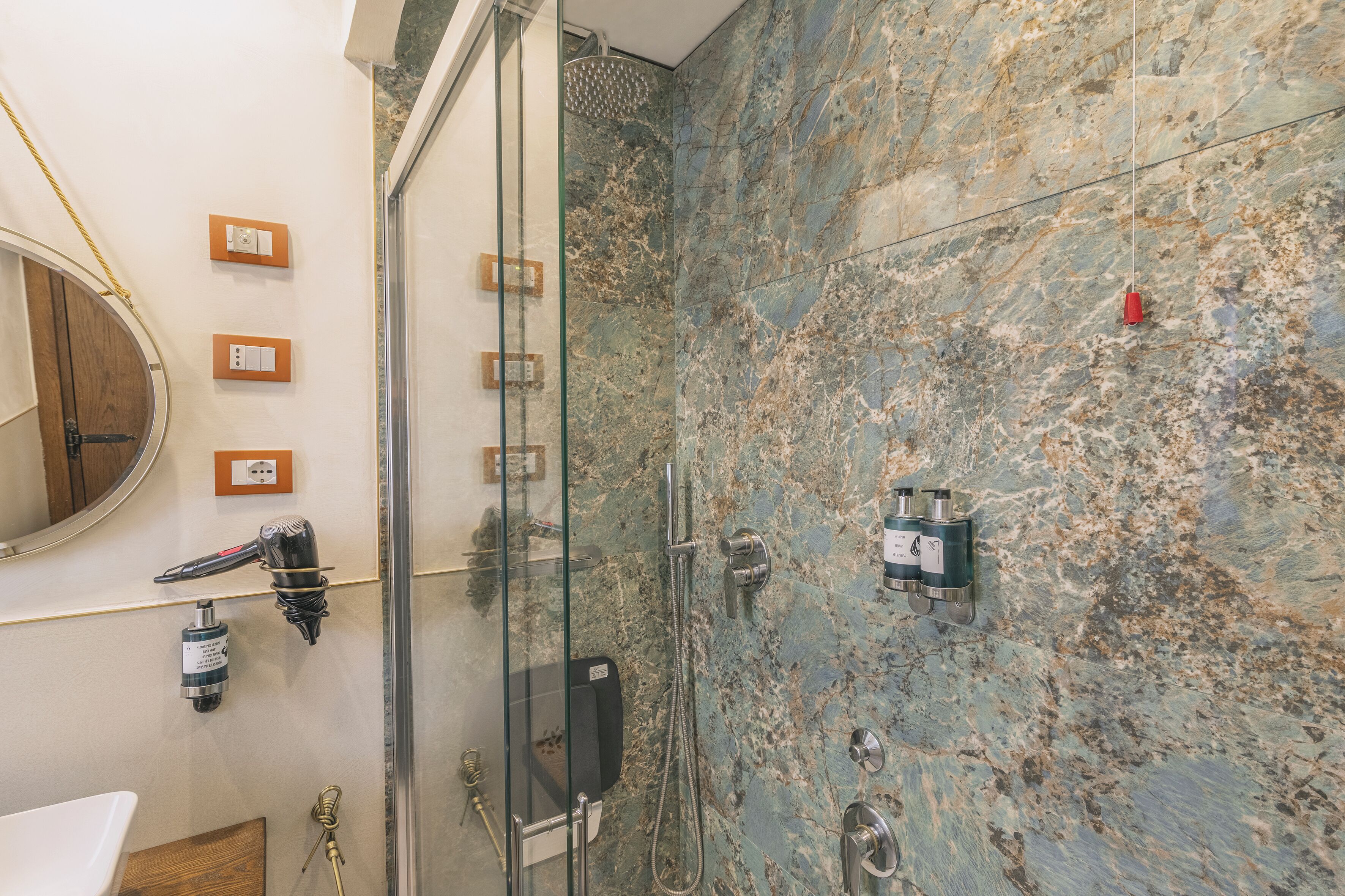 A bathroom with a glass-enclosed shower, marble walls, a round mirror, and a hairdryer mounted on the wall.