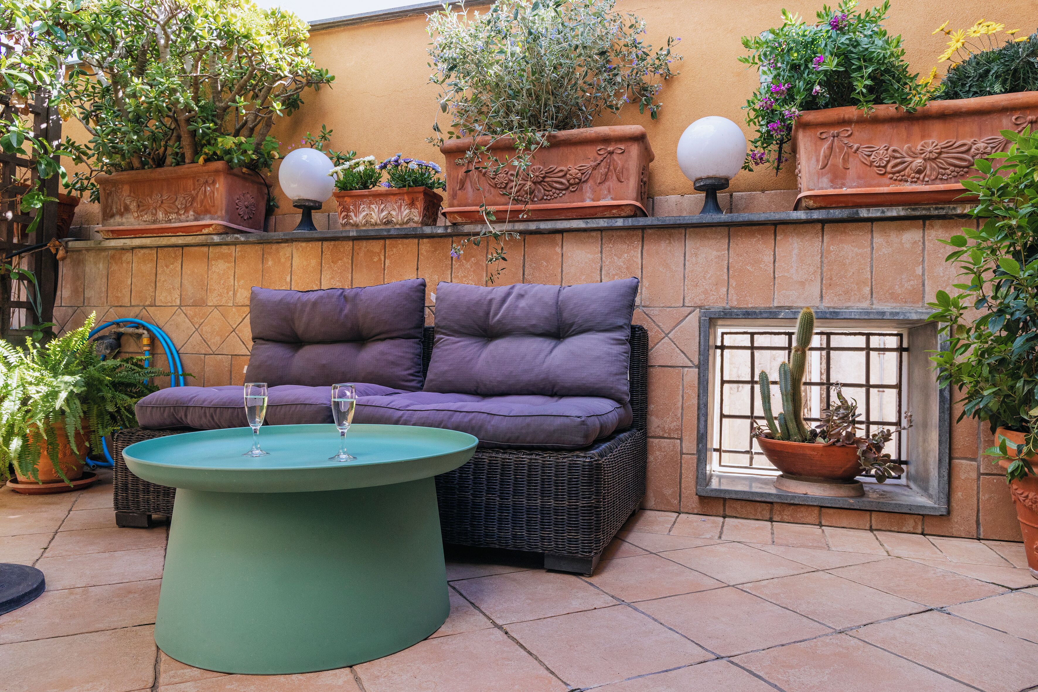 Une terrasse avec un canapé en osier, une table ronde avec deux verres et diverses plantes en pot.