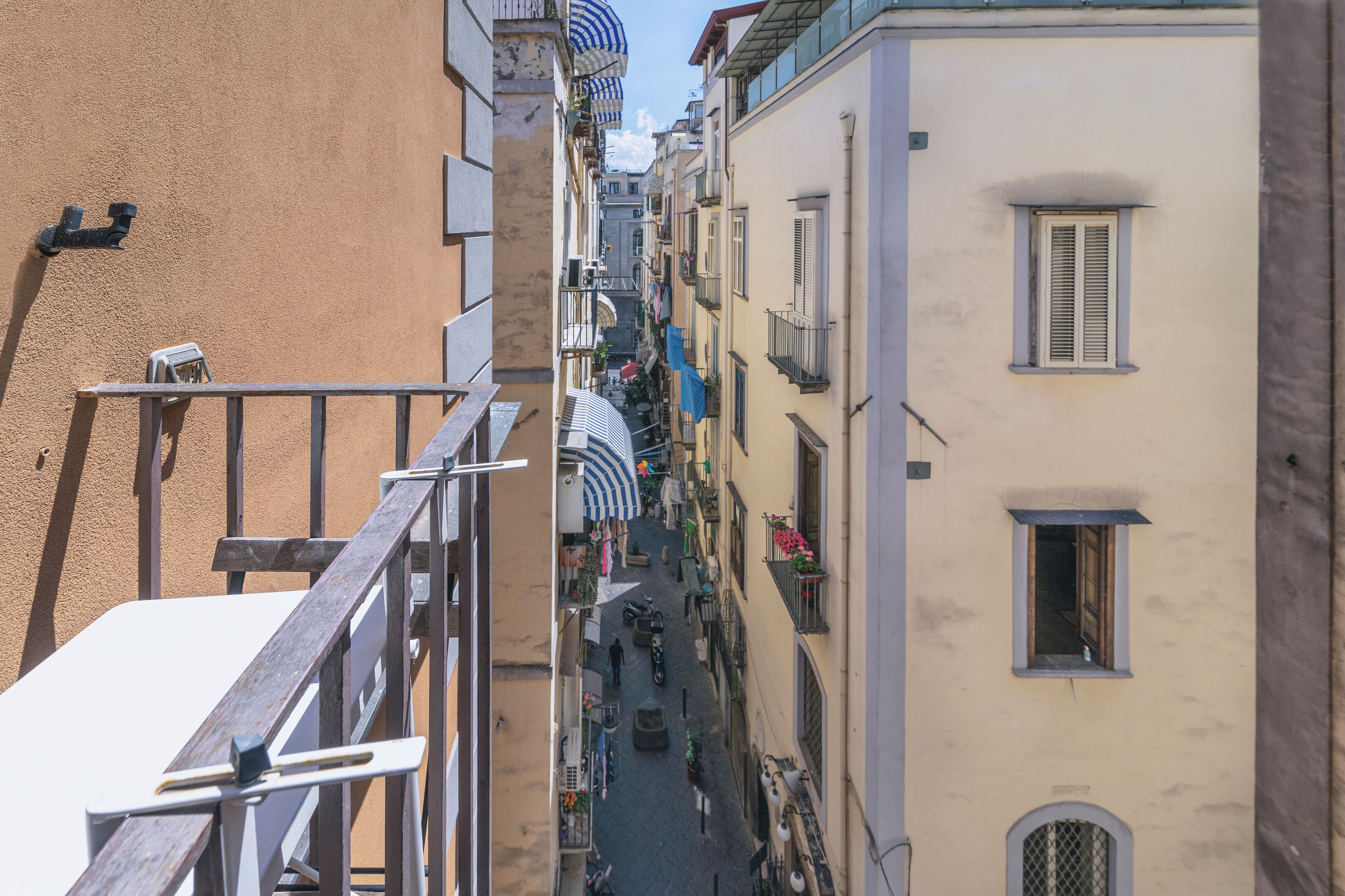 Rua estreita ladeada por edifícios com varandas, uma cerca de madeira e um toldo listrado de azul.
