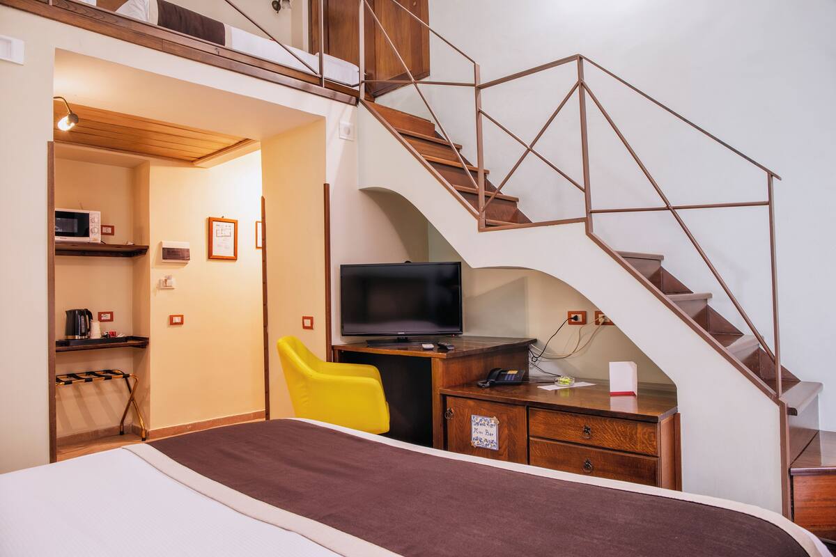 Habitación de hotel con cama, escritorio con televisor, silla amarilla, escalera de madera y pequeña cocina americana.