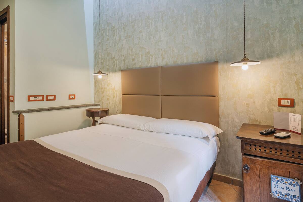Una cama bien hecha con cabecera, una mesita de noche con lámpara y cartel de minibar.