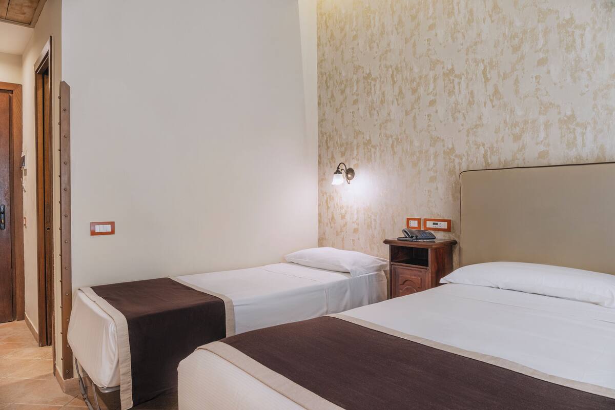 Una habitación de hotel con dos camas individuales, una mesita de noche de madera con un teléfono, y una lámpara fijada a la pared.