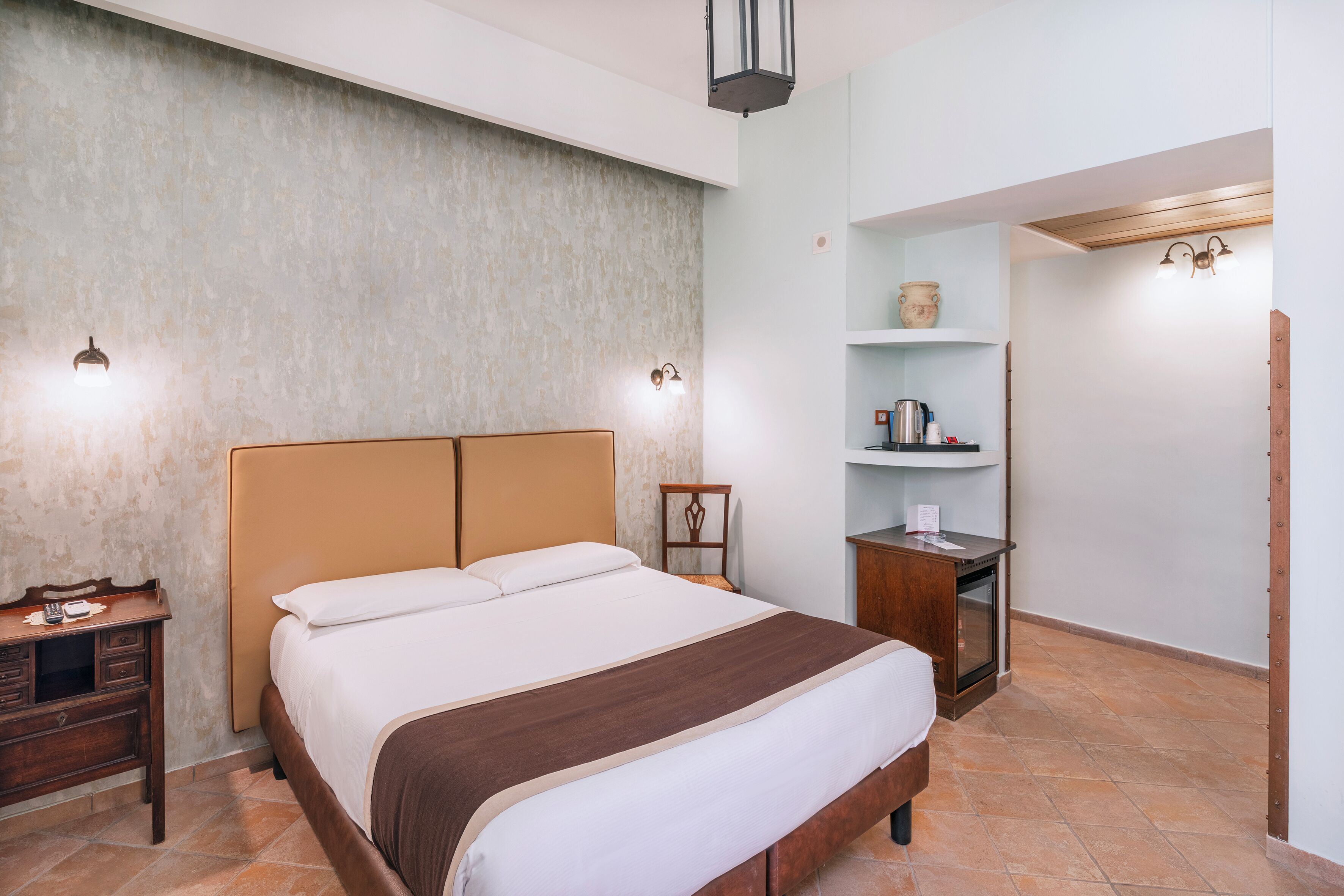 A hotel room with a large bed, a wooden nightstand, a small table, and a shelf with a vase and a kettle.