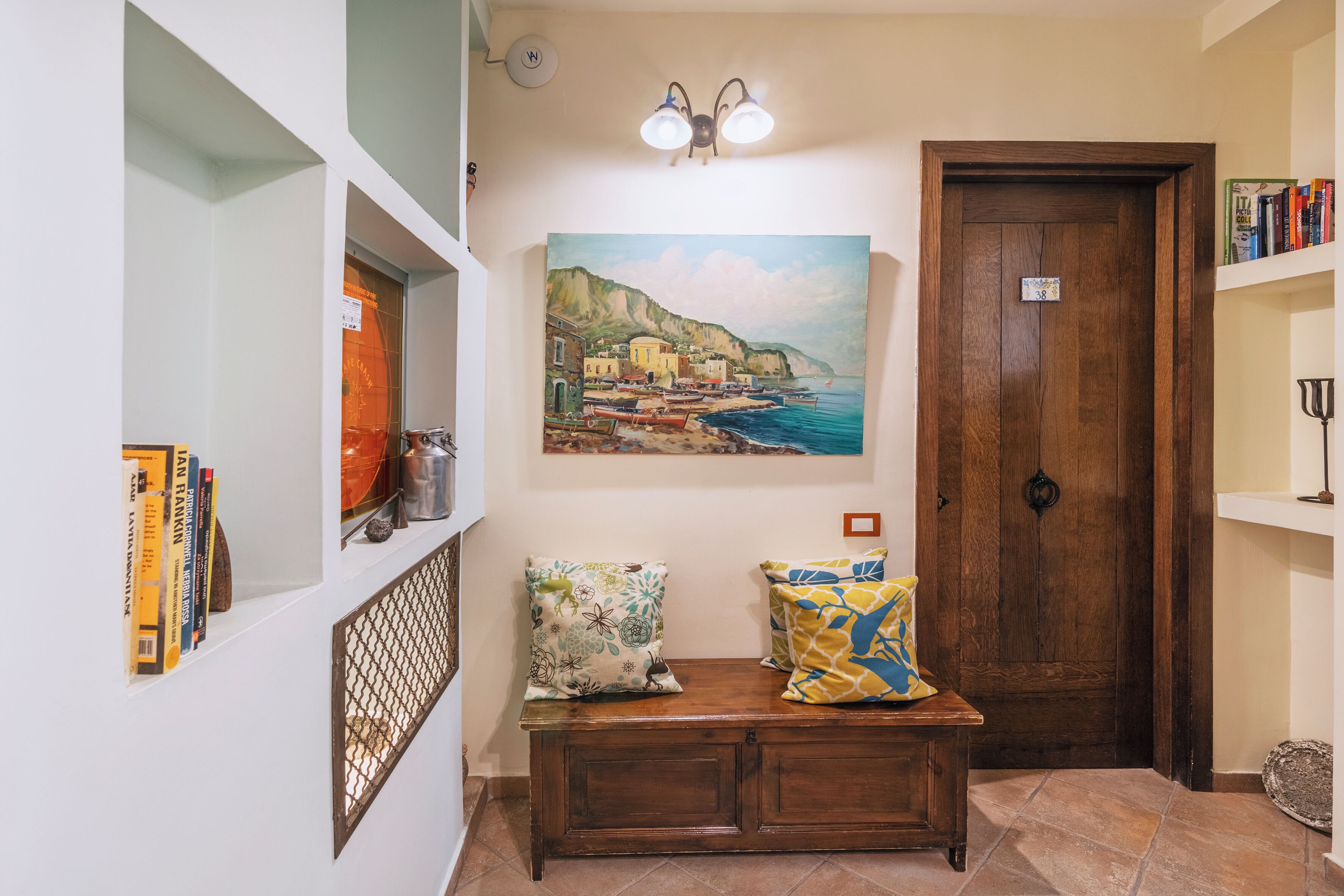 A hallway with a wooden bench, a painting of a coastal scene, a door, and a bookshelf with books.