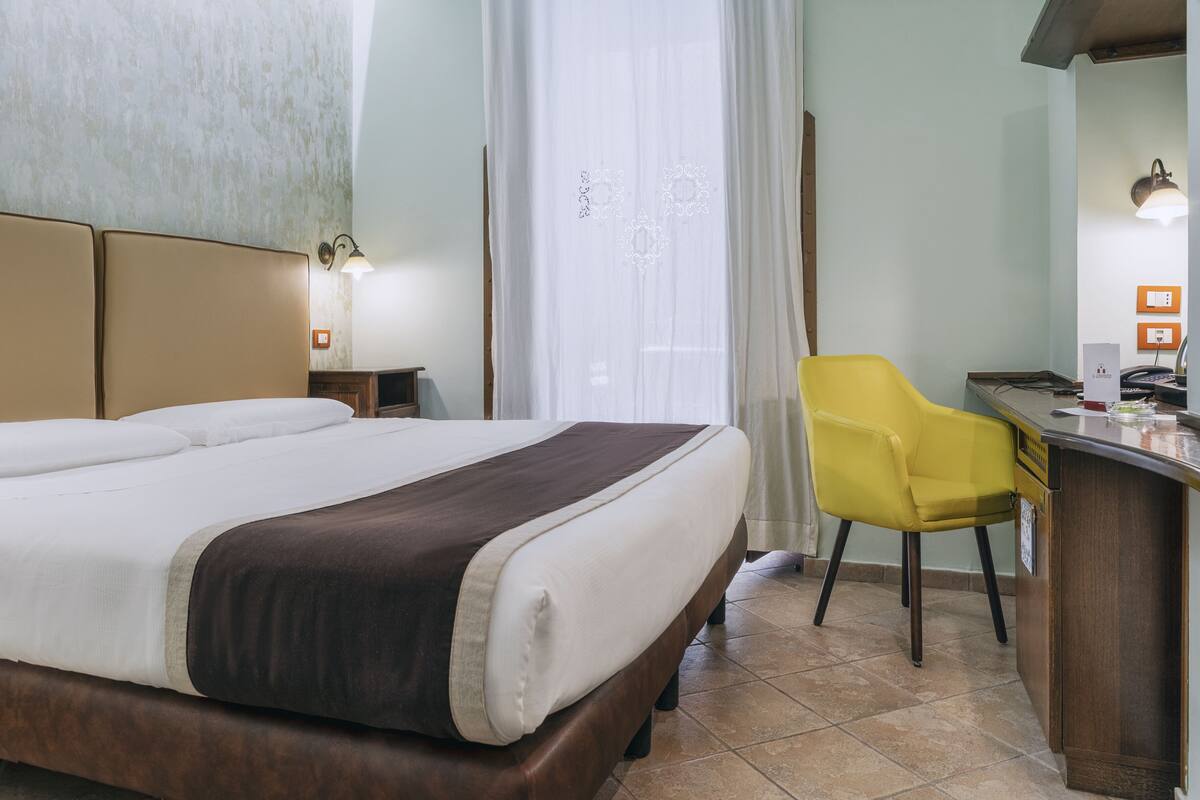 Una habitación de hotel con cama, escritorio, silla y ventana con cortinas.
