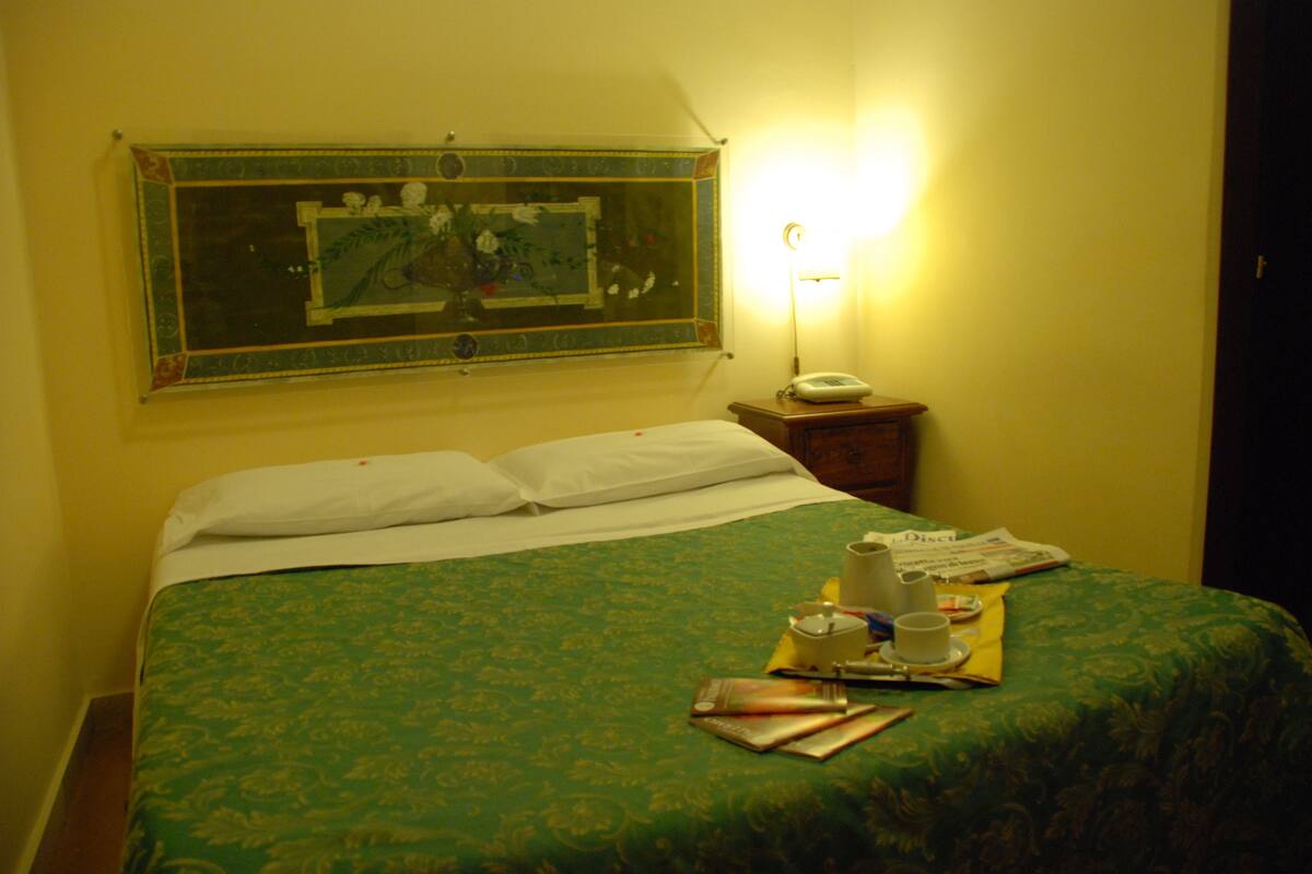 A neatly made bed with a green bedspread, a bedside table with a lamp, and a framed picture on the wall.