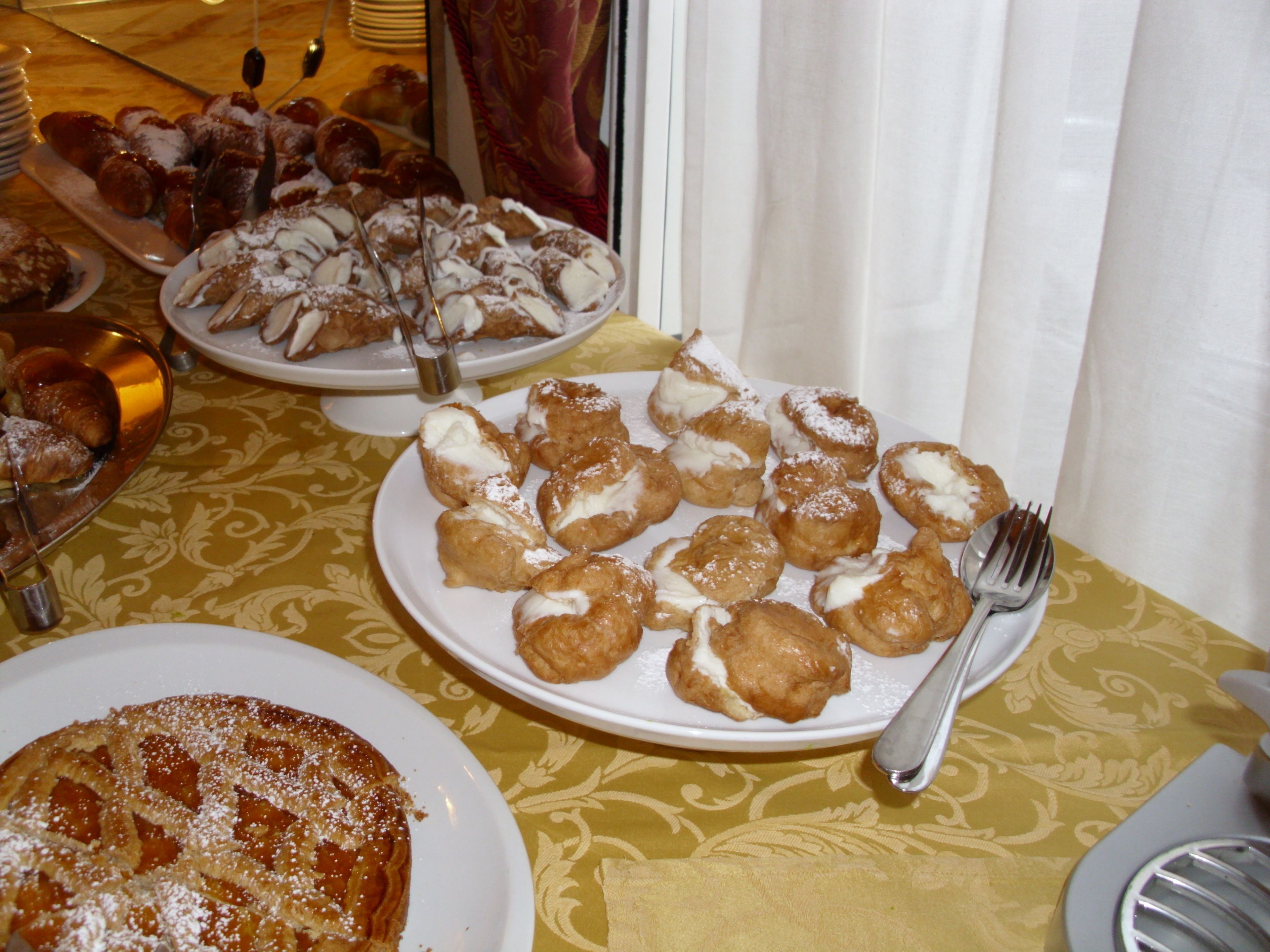 Uma mesa com pratos de pastelaria, incluindo bolas de creme e uma torta, sobre uma toalha de mesa estampada.