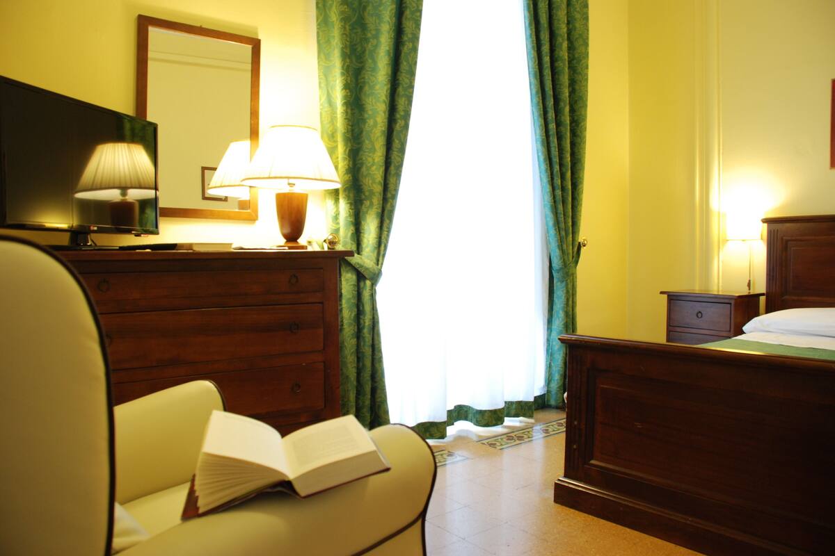A hotel room with a bed, a bedside table, a lamp, a chair, and a book on the chair.