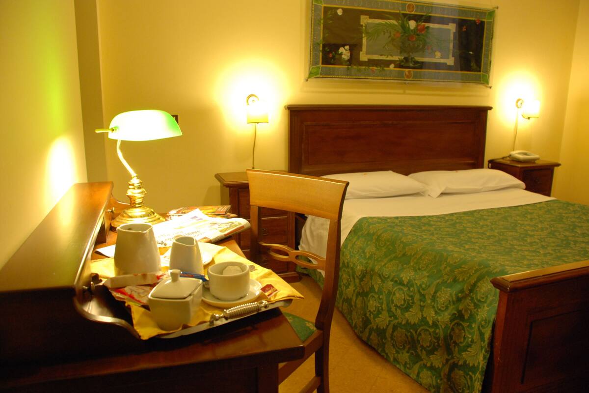 A hotel room with a bed, bedside table, lamp, and a tray with cups and a teapot.