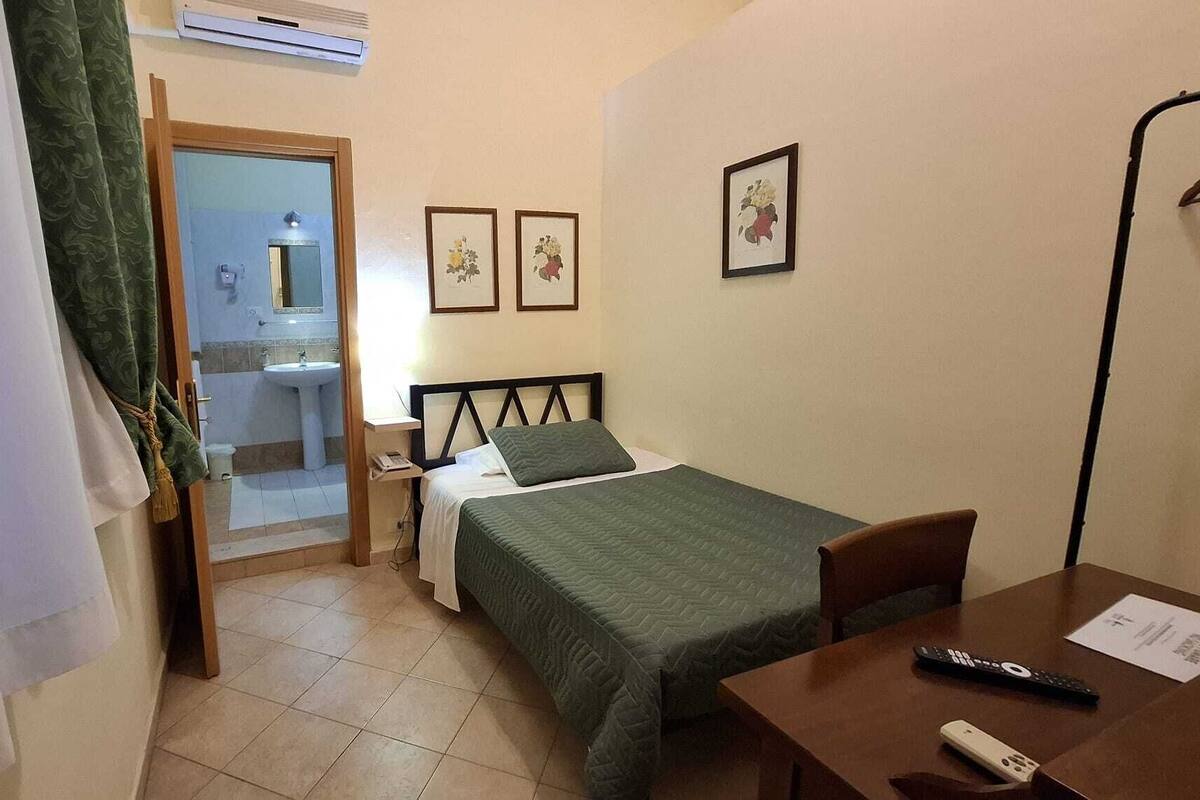 A small, tidy room with a bed, a desk, and a chair. There is a bathroom visible through an open door.