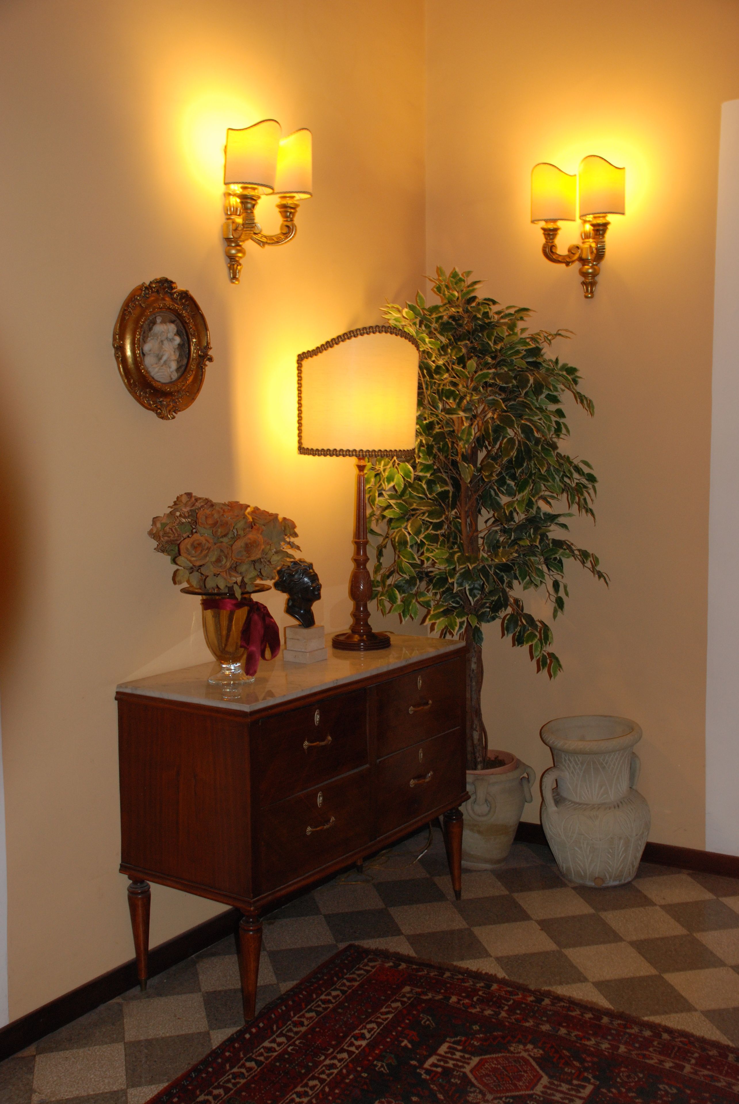 A wooden dresser with drawers, a potted plant, and a vase with flowers. There are wall sconces with lamps and a framed picture on the wall.