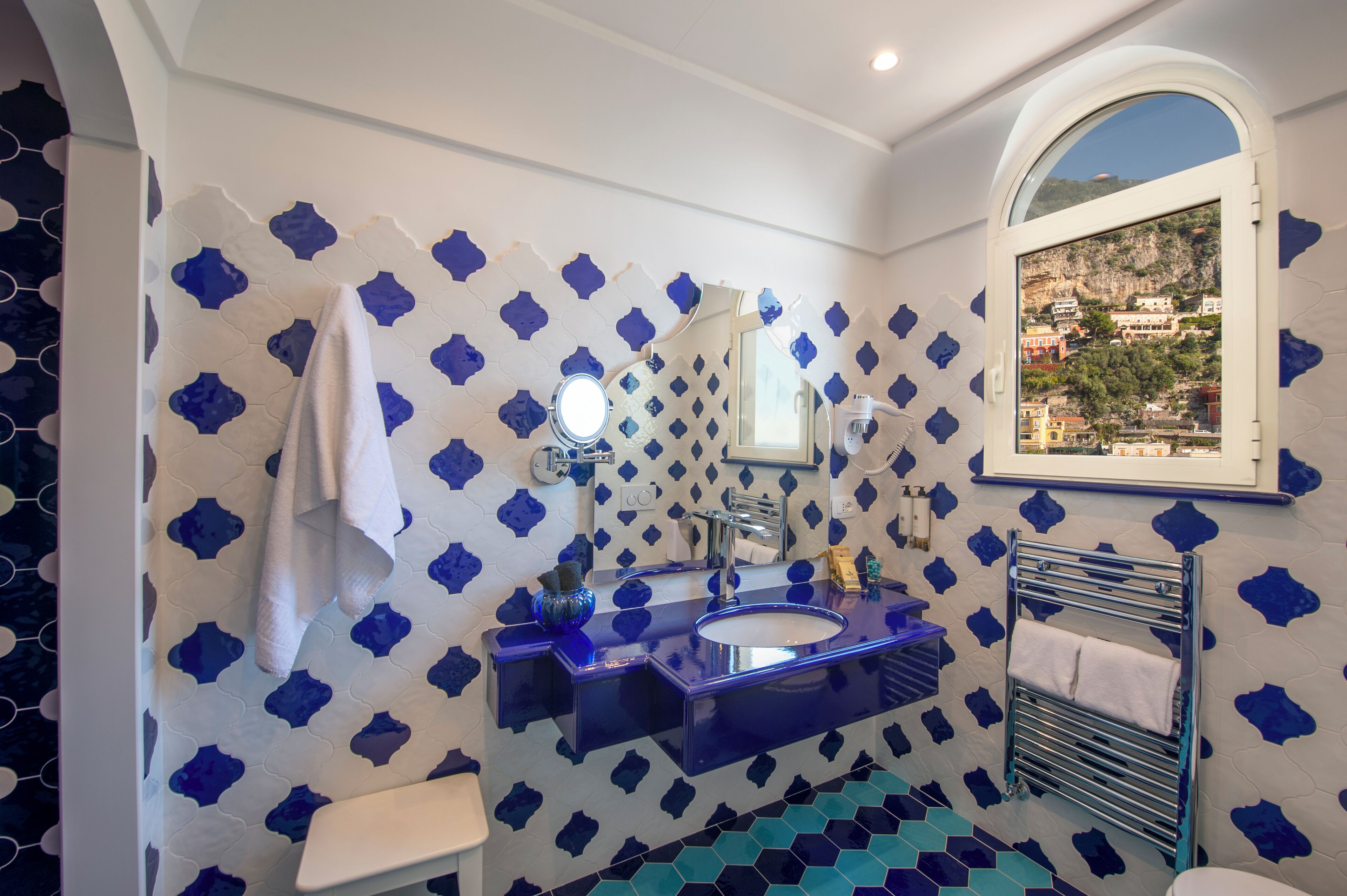 Un baño con pared de azulejos blancos y azules, un lavabo con espejo y una ventana con vista a un pueblo.