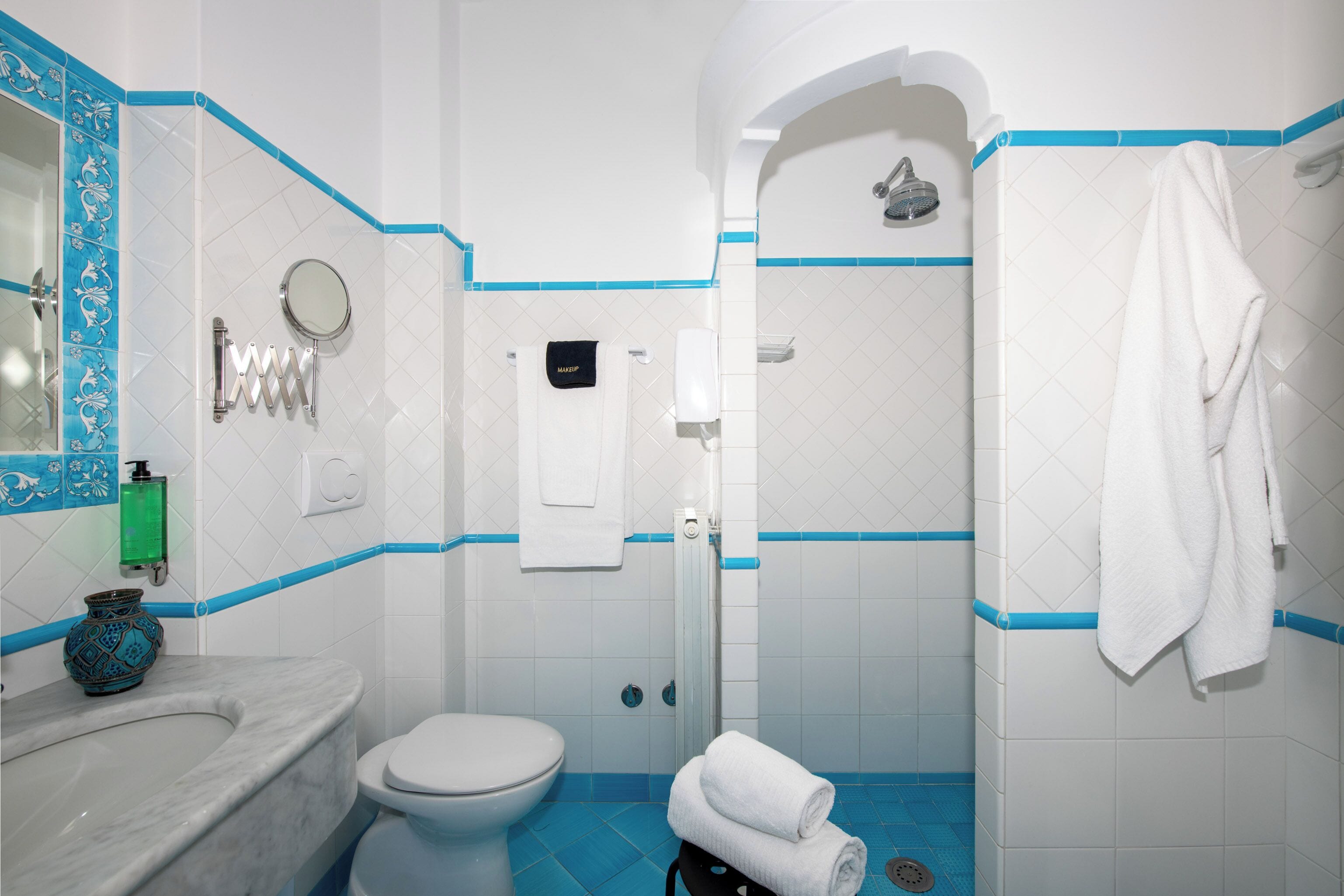 Un baño con inodoro blanco, un lavamanos con un florero azul, una ducha con piso de azulejos azules y un toallero con una toalla blanca.