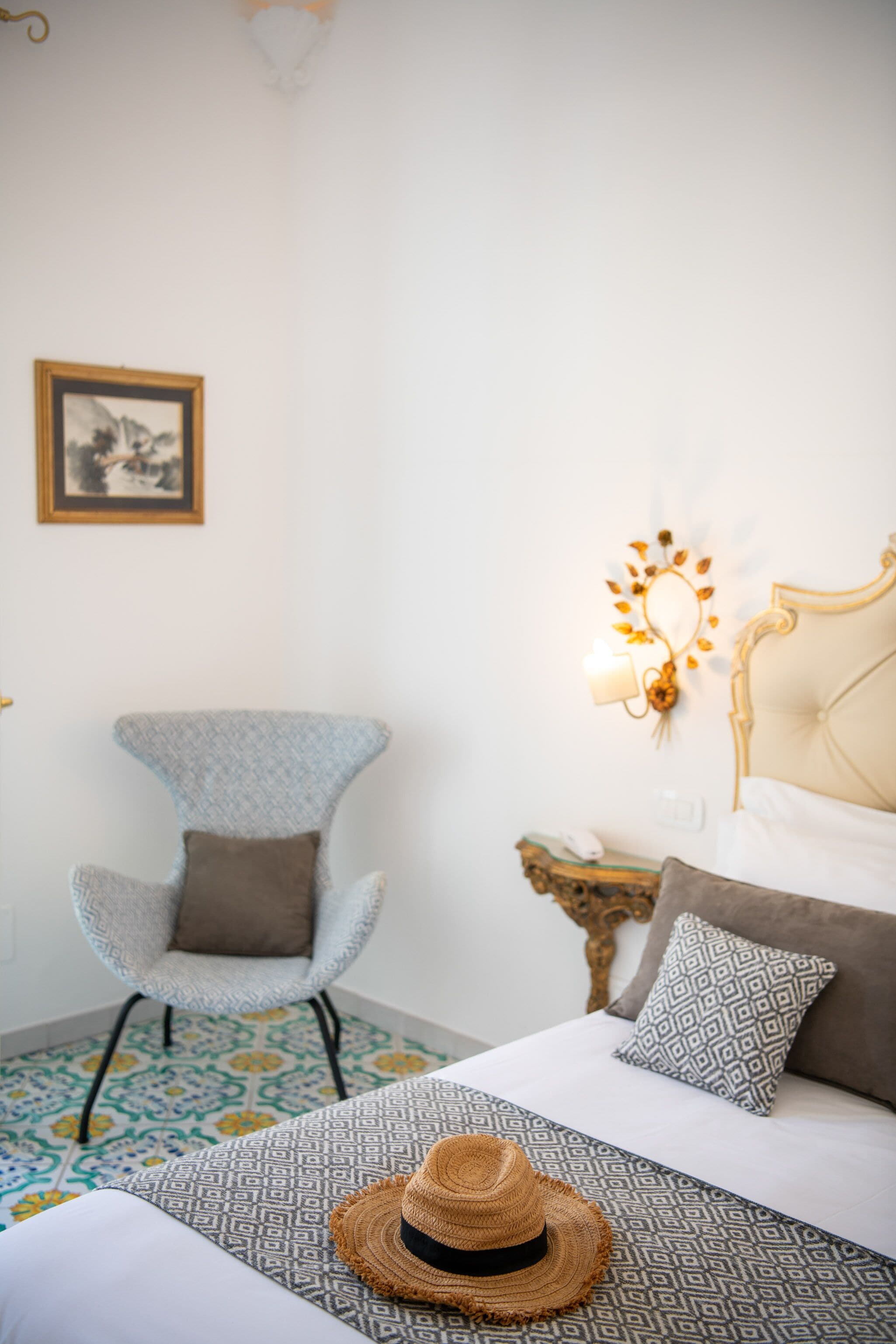 Une chambre à coucher avec une couvre-lit à motifs, un chapeau de paille posé sur le lit, une chaise, une table de chevet avec une lampe et un tableau encadré accroché au mur.