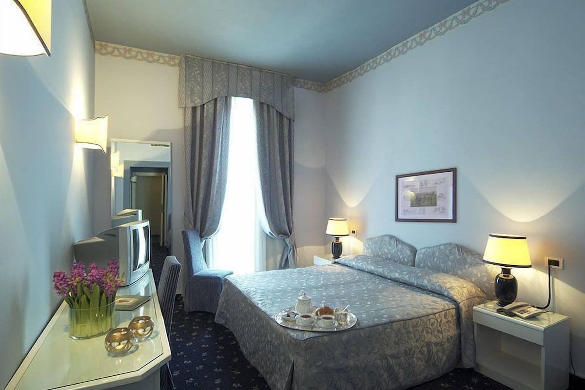 A hotel room with a bed, bedside lamps, a desk with a lamp, a TV, and a vase with flowers.
