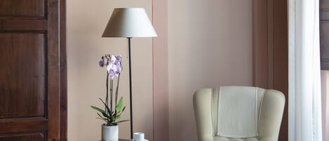 Ein Zimmer mit einem Holzkasten, einem Stuhl, einem kleinen Tisch mit einer Lampe und einem Blumentopf, sowie einem Fliesenboden.