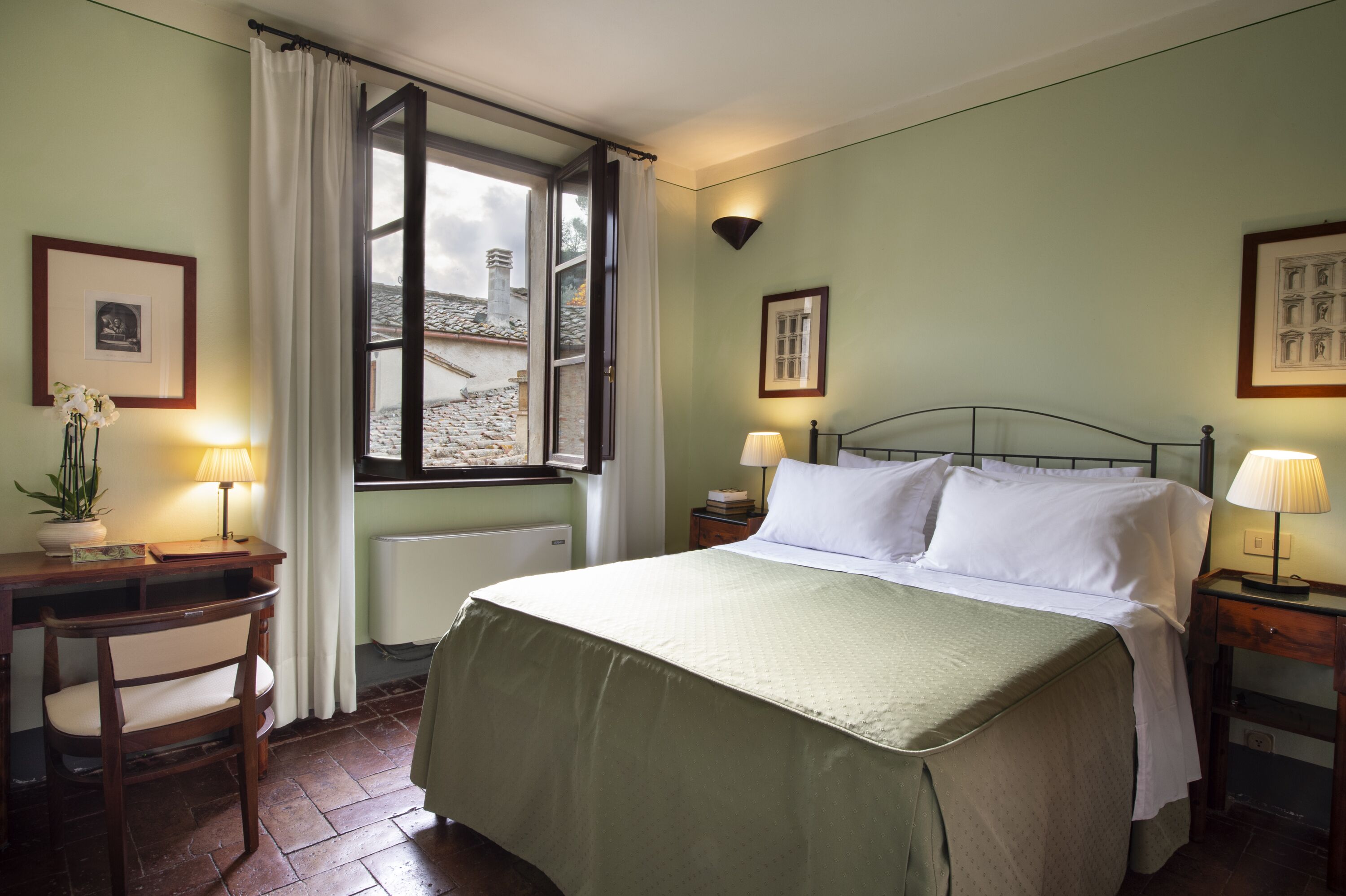 A bedroom with a bed, a desk, a chair, a window with curtains, and a radiator.