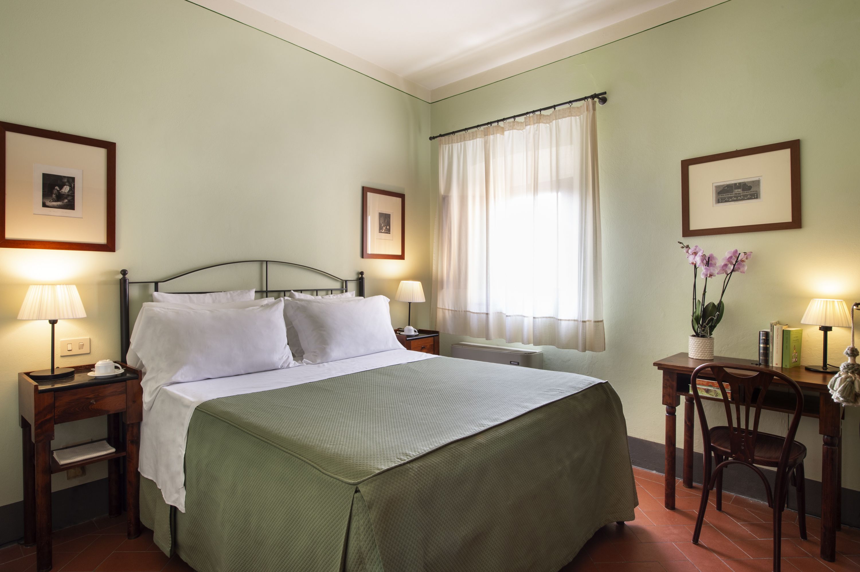 A neatly arranged hotel room with a bed, bedside tables with lamps, a small desk with a chair, and framed pictures on the walls.