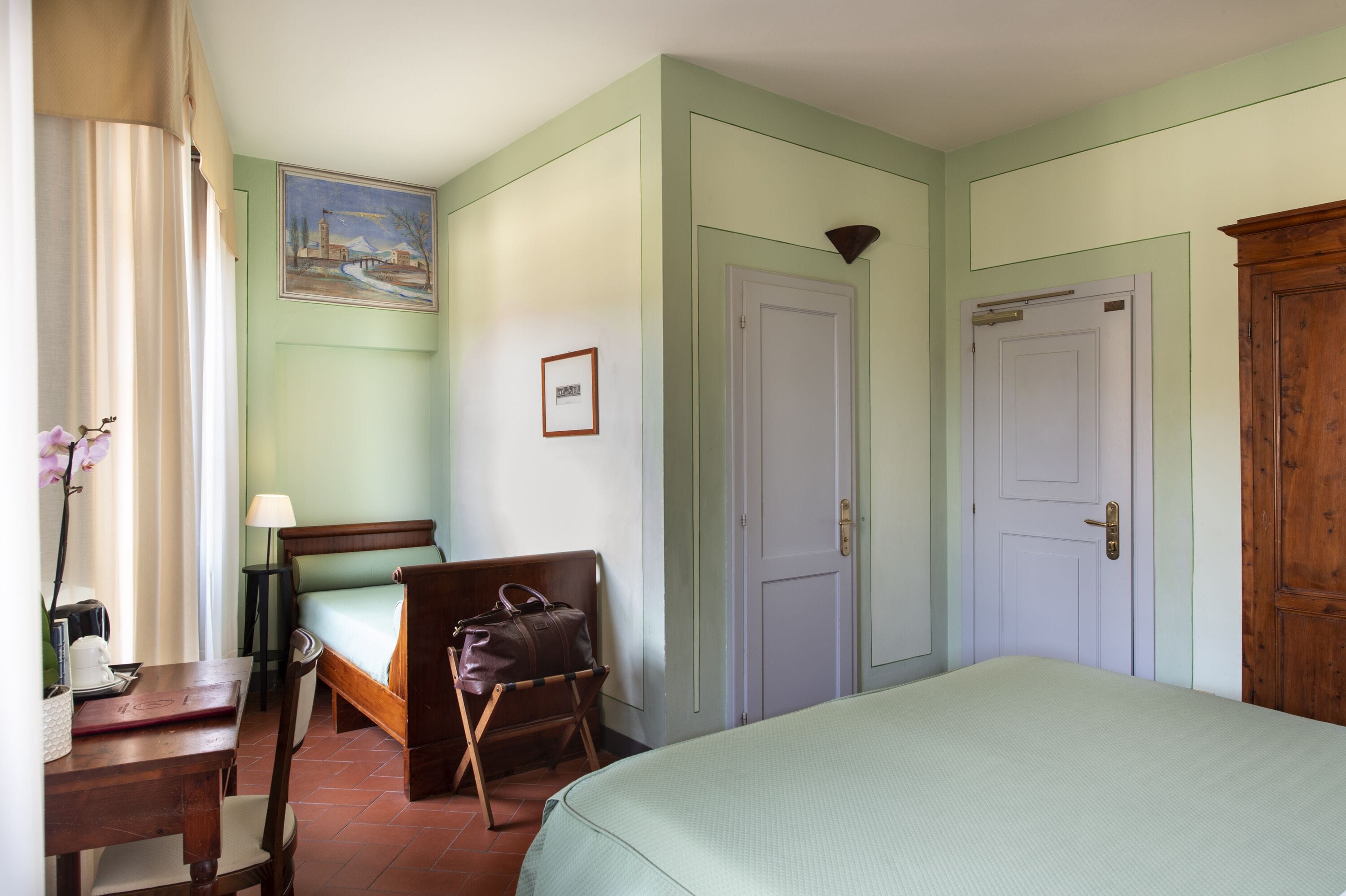 A hotel room with a bed, a desk with a lamp, a chair, a suitcase, and a painting on the wall.