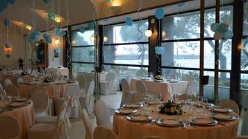 Un salón de banquetes con mesas redondas dispuestas para cenar, sillas blancas y ventanas amplias que dan a vistas del exterior.