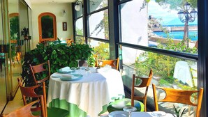 Un comedor con mesas dispuestas para dos, ventanas amplias que ofrecen vista al mar y follaje, y una planta en maceta en el centro.