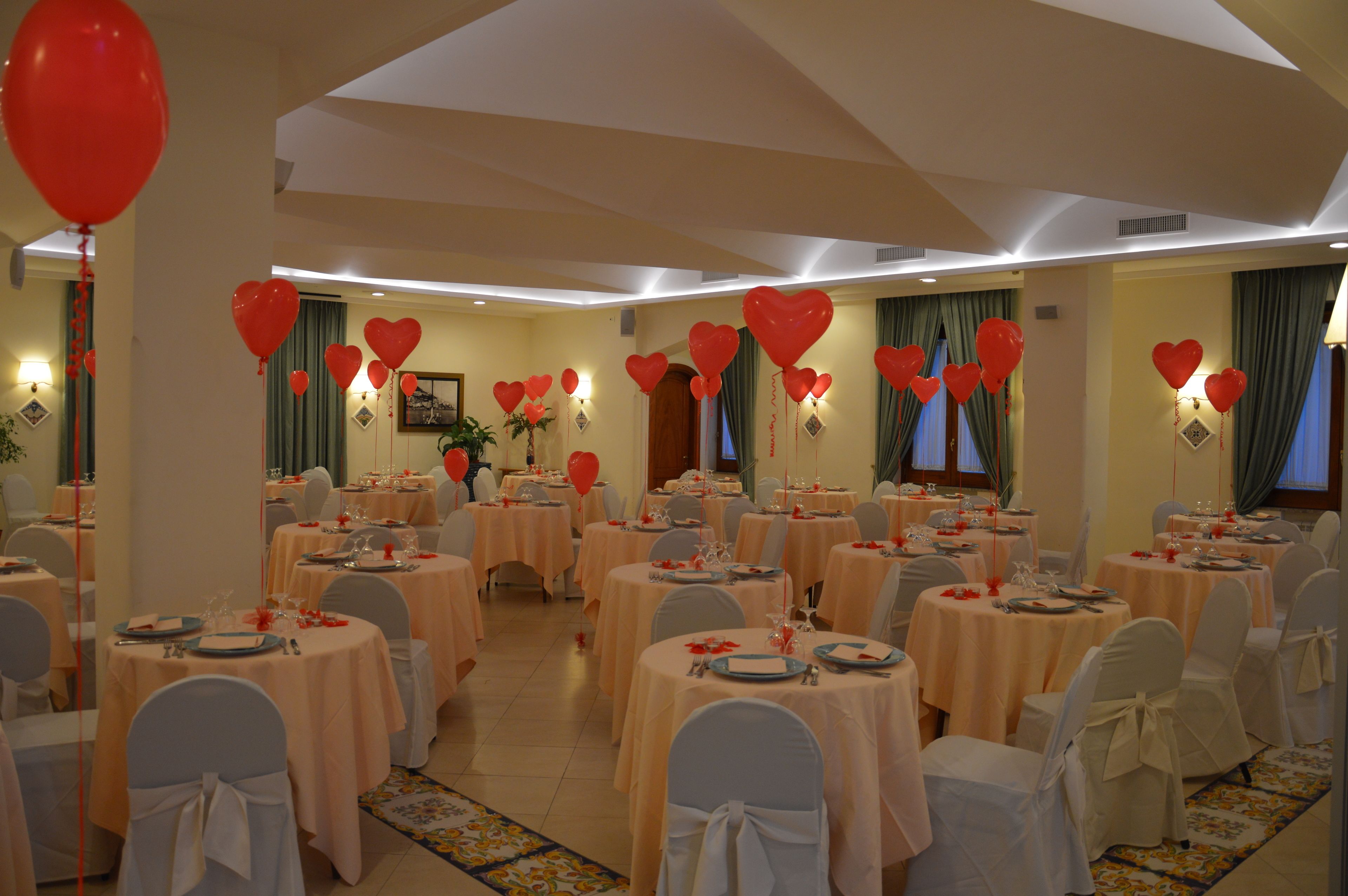 A banquet hall with round tables, white chairs, and red heart-shaped balloons.