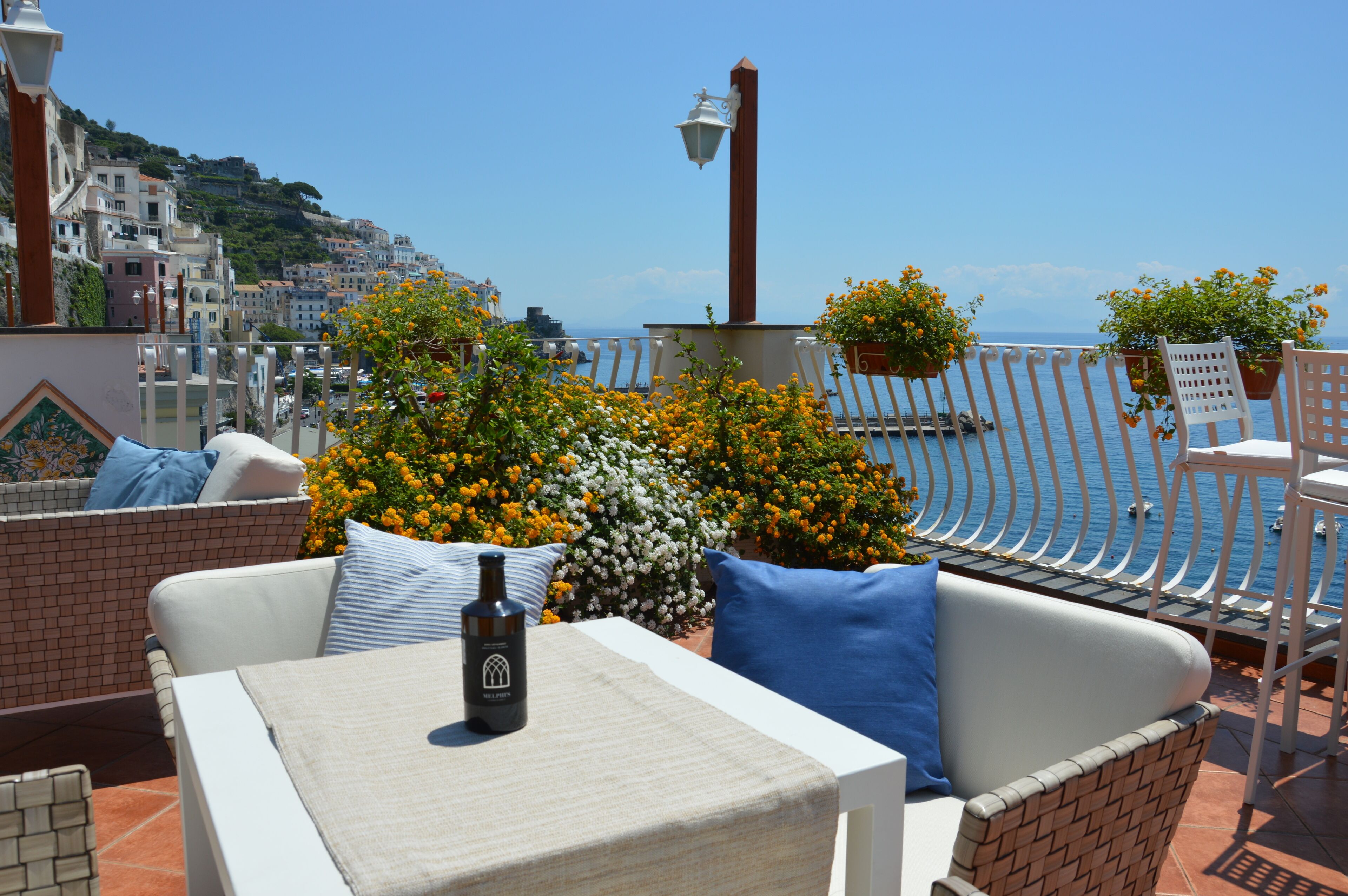 Eine Terrasse mit weißem Korbmöbel, einem Tisch mit einer Flasche und Kübelpflanzen mit orangefarbenen Blüten.