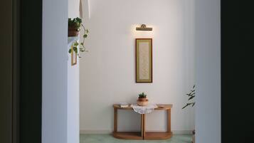 Un pasillo estrecho con un arco blanco que da a una habitación con un banco de madera, una mesita con una planta y un cuadro en la pared.