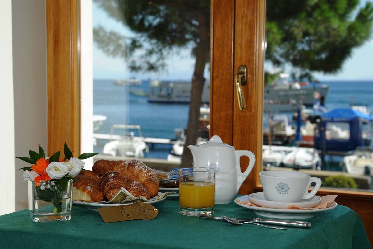 A breakfast table with croissants, a glass of orange juice, a teapot, and a cup, set against a backdrop of a marina with boats and trees.