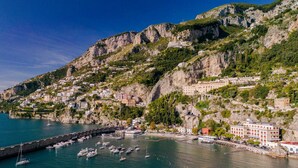 Un pueblo costero con edificios en una ladera, un puerto deportivo con barcos y un acantilado rocoso.