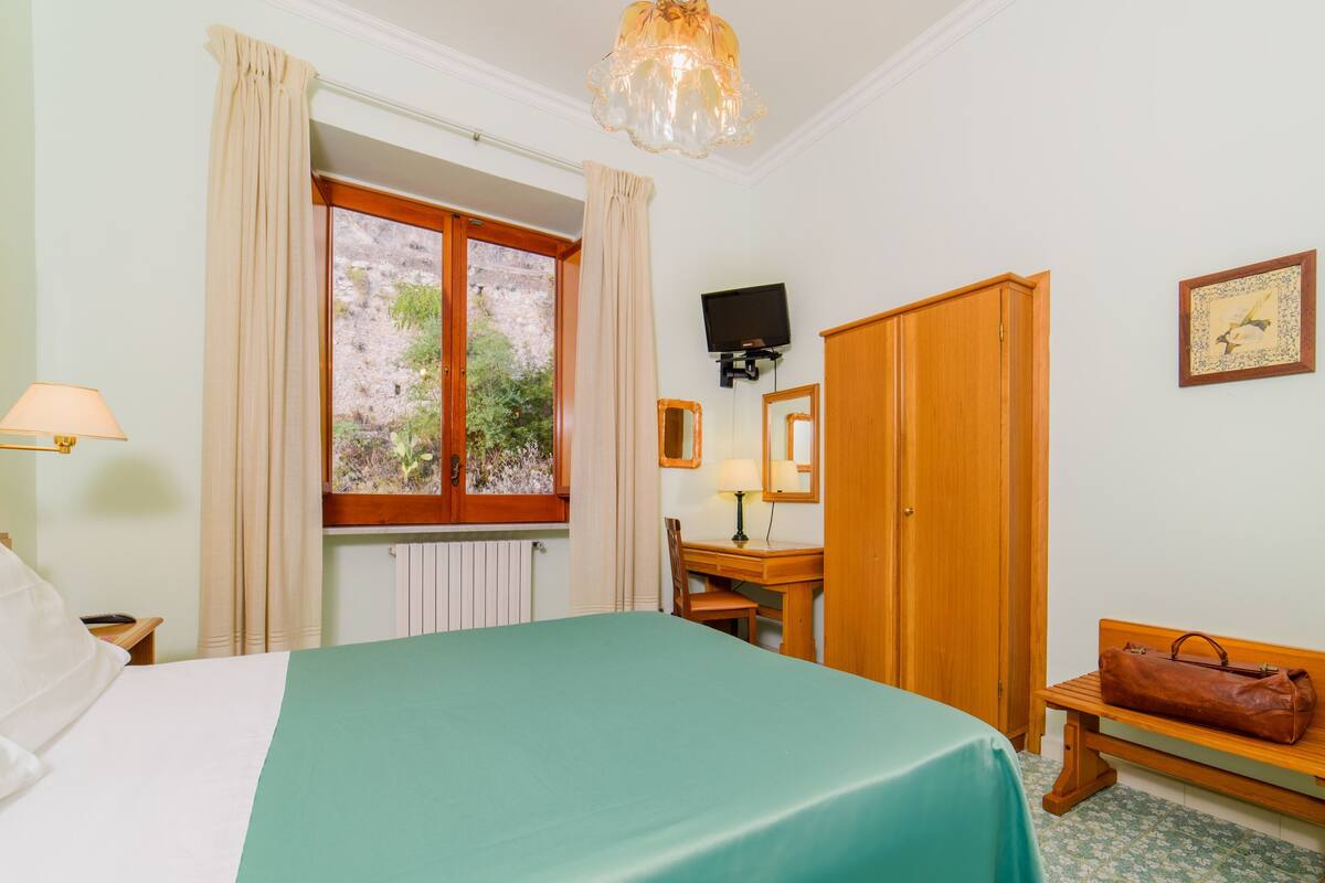 A hotel room with a bed, a desk, a television, a wardrobe, and a bench.